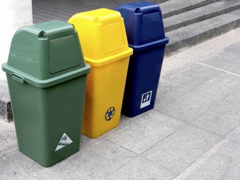 Three recycling bins are positioned on a paved sidewalk surface, arranged side by side with their lids closed. The first bin on the left is green with a smooth plastic texture, featuring a triangular recycling symbol and a small white label near its base. The middle bin is bright yellow, also with a smooth surface, displaying a circular recycling icon in blue on its front panel. The third bin on the right is deep blue, slightly taller, with a white label or icon on its side. The bins are located outdoors, adjacent to a building with a step or ramp leading up to it, and are set on a clean, grey stone pavement. The surrounding environment suggests a residential or commercial area in London, possibly near Regents Park, supporting local rubbish collection and waste management services. The lighting indicates a bright, clear day, emphasizing the vibrant colours of the bins and the neat condition of the paved area. Garden Clearance Regents Park likely supplies these bins as part of their rubbish removal services, facilitating proper waste separation and disposal within the local community.