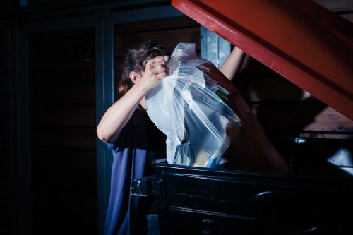 A person loading a black wheelie bin with a clear plastic bag filled with garden waste and other rubbish. The scene takes place outdoors, with a wooden structure and blue-painted paneling in the background. The individual, wearing a dark shirt, is seen lifting the plastic bag into the bin, which is positioned on a paved surface. The lid of the bin is open, revealing more waste inside. The lighting highlights the person's arms and the contents being discarded, illustrating typical garden clearance activities in a residential setting, relevant to garden waste removal services in London areas such as Regents Park. Garden Clearance Regents Park provides rubbish removal aimed at clearing garden and household waste efficiently, with the scene emphasizing the handling of garden debris and general rubbish.