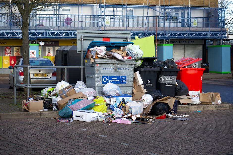 A middle-aged man with dark hair, wearing a black T-shirt with white text and black pants, is seen bending over to dispose of waste into a large, stainless steel public litter bin situated on a paved