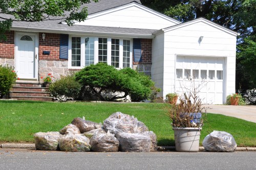 A residential front yard in Regents Park featuring a well-maintained lawn with a slight slope leading to a brick and white-panelled house. In the foreground, several large plastic garbage bags filled with yard waste are lined up along the curb, some partially transparent revealing leaves and garden debris inside. To the right, there is a small potted plant with a few dried branches emerging from it, placed on the pavement. Behind the bags, a low green shrub and a larger, well-pruned bush with dark green foliage partially obscure a set of brick steps leading to the front door, which is white with a small window and a black mailbox mounted beside it. The house has a prominent bay window with multiple panes and a white frame, complemented by blue shutters on the sides. The scene is brightly lit, indicating a clear day, with shadows cast on the ground. The overall setting suggests a garden clearance activity in progress, typical of a rubbish removal service in the local area, with Garden Clearance Regents Park managing disposal of yard waste and garden rubbish.