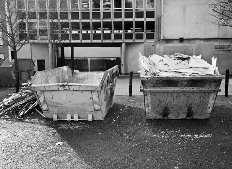 Two large industrial skips positioned on a paved surface in front of a modern building with a grid-patterned facade. The left skip contains broken concrete, bricks, and rubble with rough, uneven textures and light grey tones. The right skip is filled with a pile of white plasterboard sheets and various debris, with the sheets stacked haphazardly, some leaning or slightly bent. Surrounding the skips are small scattered debris, including bits of plaster and dust. The background shows a concrete wall and window panels of the building, with a few leafless trees and metal bollards nearby. The scene suggests a location where rubbish removal and clearance services, such as those provided by Garden Clearance Regents Park, are carried out, reflecting typical garden or construction waste disposal in the London area.