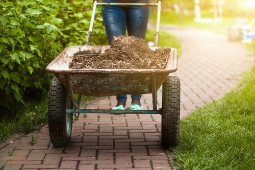 A person wearing blue jeans pushes a wheelbarrow filled with dark, moist soil along a paved pathway in a garden. The wheelbarrow is made of metal with a rust-colored finish and has black rubber tires. The pathway is bordered by green grass and dense shrubs on the left side, with a neatly maintained garden area. In the background, the pathway continues into a sunny outdoor setting, indicating a residential or landscaped environment. This scene illustrates a garden clearance activity, typical for rubbish removal services in Regents Park, with Garden Clearance Regents Park specialising in clearing soil, garden waste, and debris from outdoor spaces.