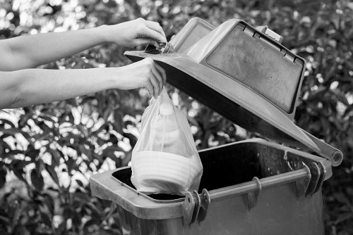 A person loading a plastic bag of discarded ceramic bowls into a large black wheelie bin with an open grey lid, positioned outdoors on a paved surface. The image shows the person's hands and forearms, with the bin placed against a backdrop of leafy shrubs or trees. The bin appears to be made of durable plastic with metal hinges and orange latches. The outdoor environment is well-lit, possibly by natural daylight, highlighting the textures of the plastic bin and the smooth surface of the ceramic bowls. The scene is set in a garden or driveway area within an urban locality, consistent with rubbish removal services in the Regents Park area. The focus is on waste disposal and rubbish collection, aligning with the services provided by Garden Clearance Regents Park.